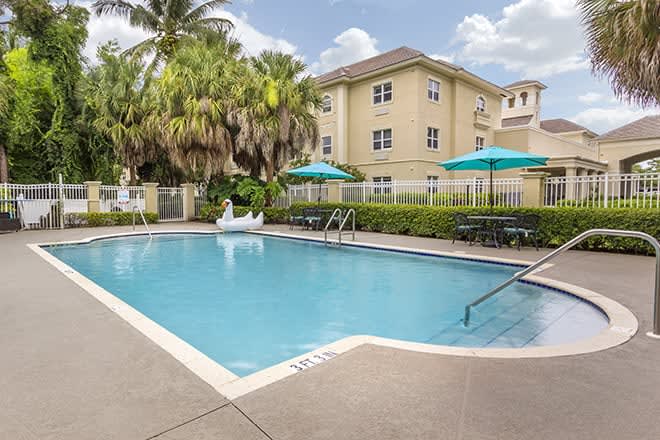 A tranquil outdoor swimming pool surrounded by palm trees and a building, featuring sun umbrellas for relaxation on a sunny day.