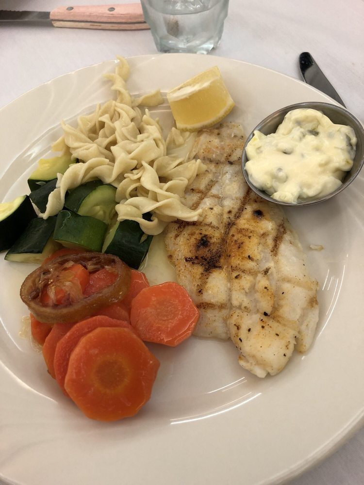 A plate of grilled fish served with sliced carrots, zucchini, cabbage, a lemon wedge, and tartar sauce.