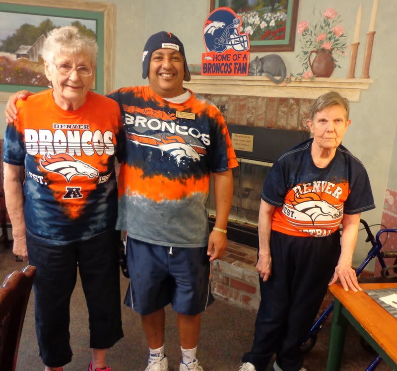 Three people wearing Broncos shirts pose for a group photo in a living room.