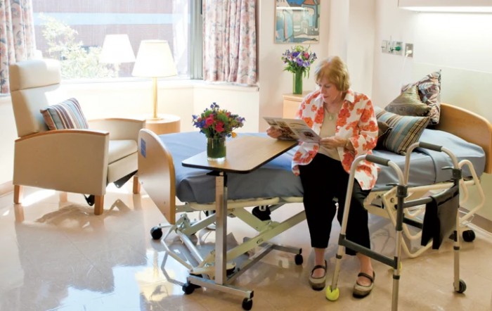 A patient sitting in a hospital room, reading next to a bed with a bouquet of flowers on the table.