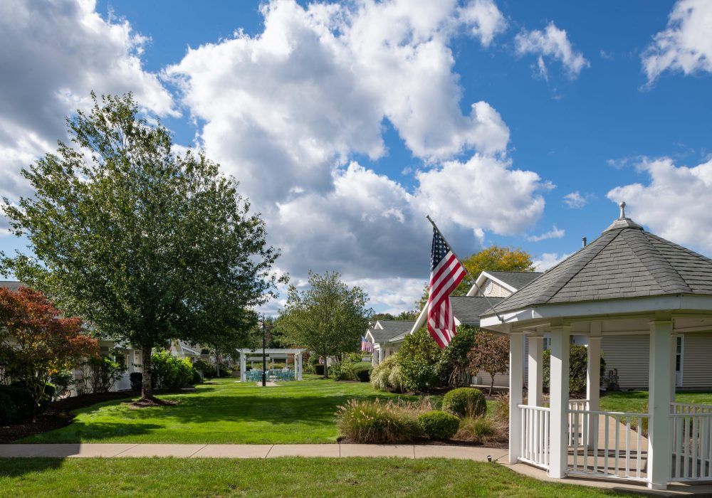 A peaceful suburban scene with a gazebo, American flag, and lush greenery under a partly cloudy sky.