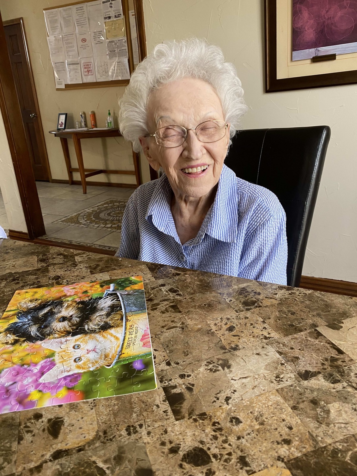 An elderly woman smiling while sitting at a table with a puzzle.
