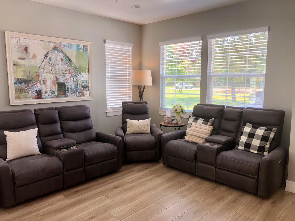 A cozy living room featuring recliner chairs and a sofa, with decorative pillows and a floor lamp by the windows. Artwork adorns the wall, creating a comfortable and inviting space.