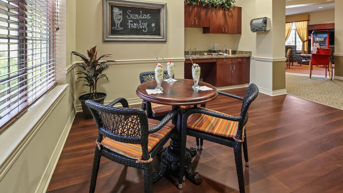 A cozy indoor seating area with a round table and woven chairs, decorated with plants and a chalkboard.
