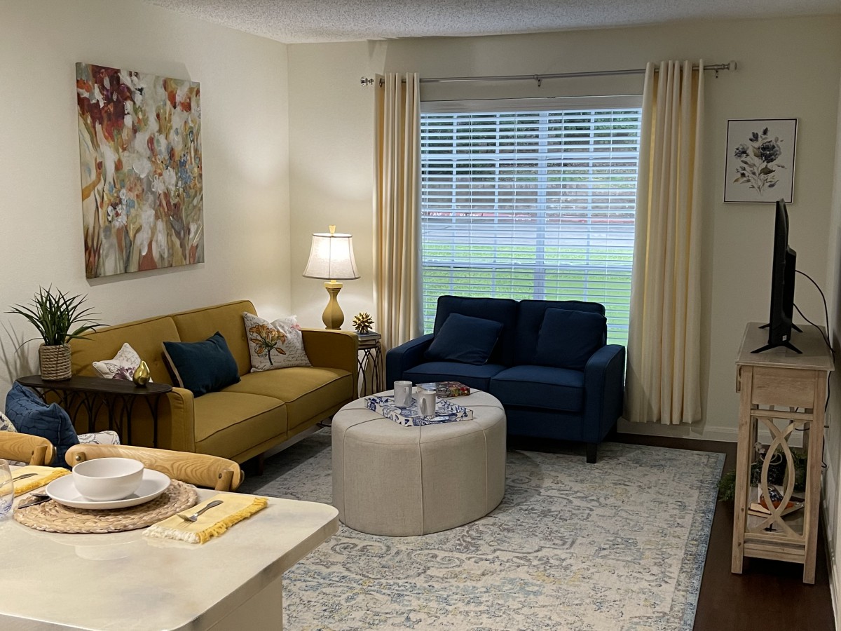 A cozy living room with a mix of modern and classic furniture, featuring a yellow and a blue sofa, an ottoman-style coffee table, and soft lighting from a table lamp.