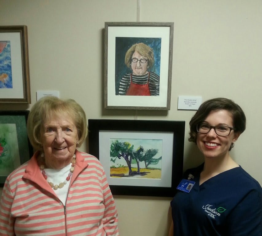 Two women stand in front of artwork displayed on a wall, including a portrait and a landscape painting.