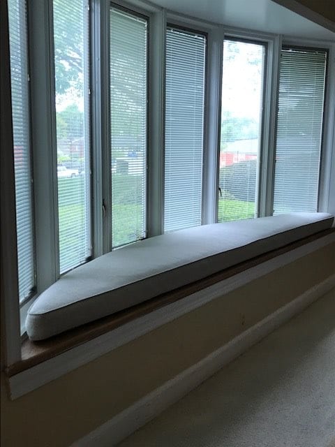 A cozy window seat with a cushion, framed by large windows and blinds, offering ample natural light.