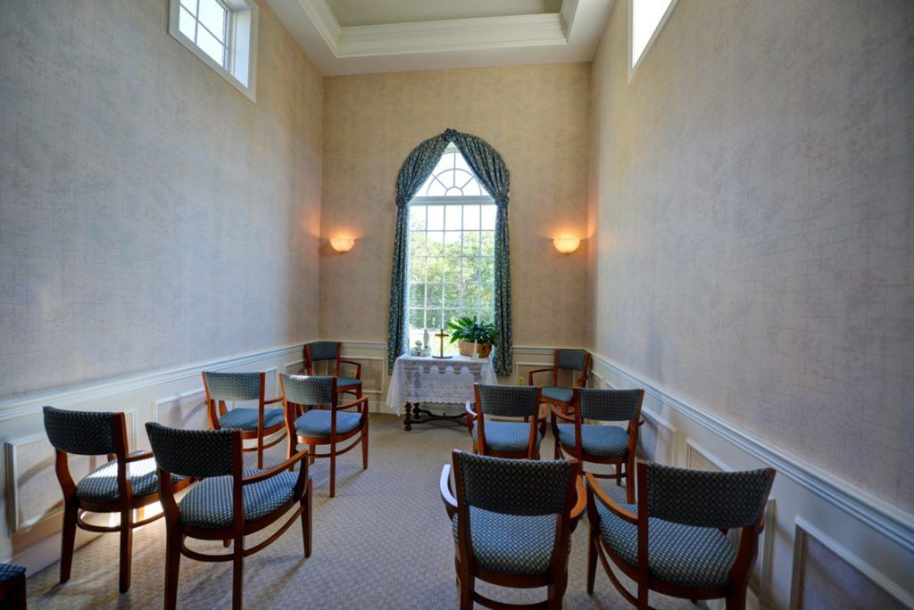 A small, elegantly designed room with rows of chairs facing a large window adorned with curtains, illuminated by natural light and wall lamps.