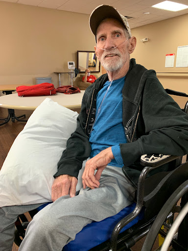 An elderly man sitting in a wheelchair, smiling and looking comfortable in a casual setting.