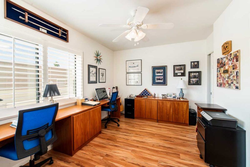 A home office setup featuring wooden desks, chairs, and a computer, with wall decor and a window providing natural light.