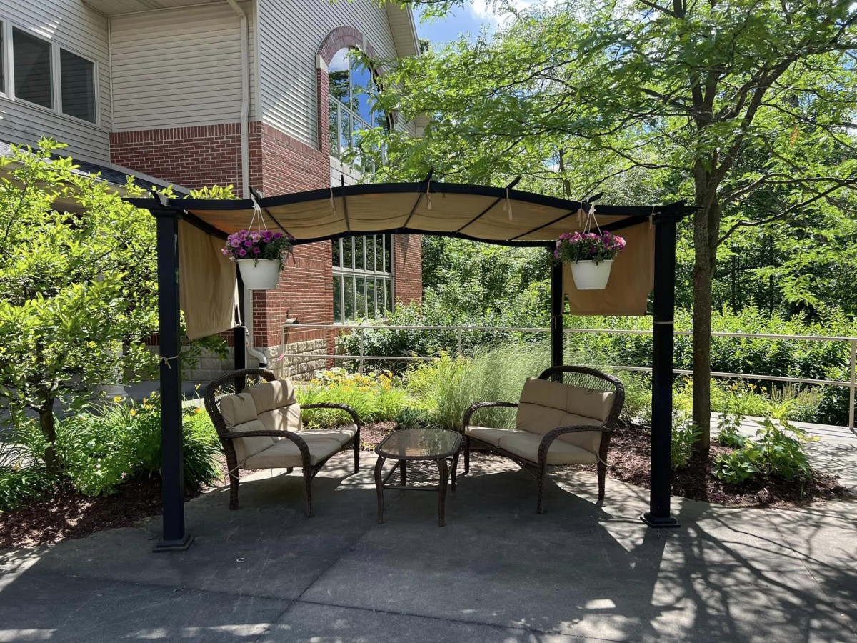A cozy outdoor gazebo with chairs and a table, surrounded by greenery and hanging plants.