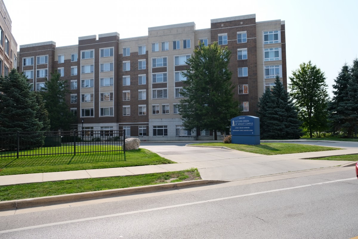 A multi-story residential apartment building with a sign and surrounding trees along a road.