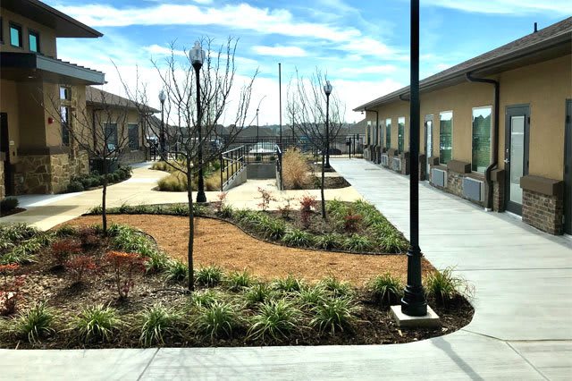 A landscaped courtyard featuring pathways and buildings, with lamp posts lining the area under a clear blue sky.