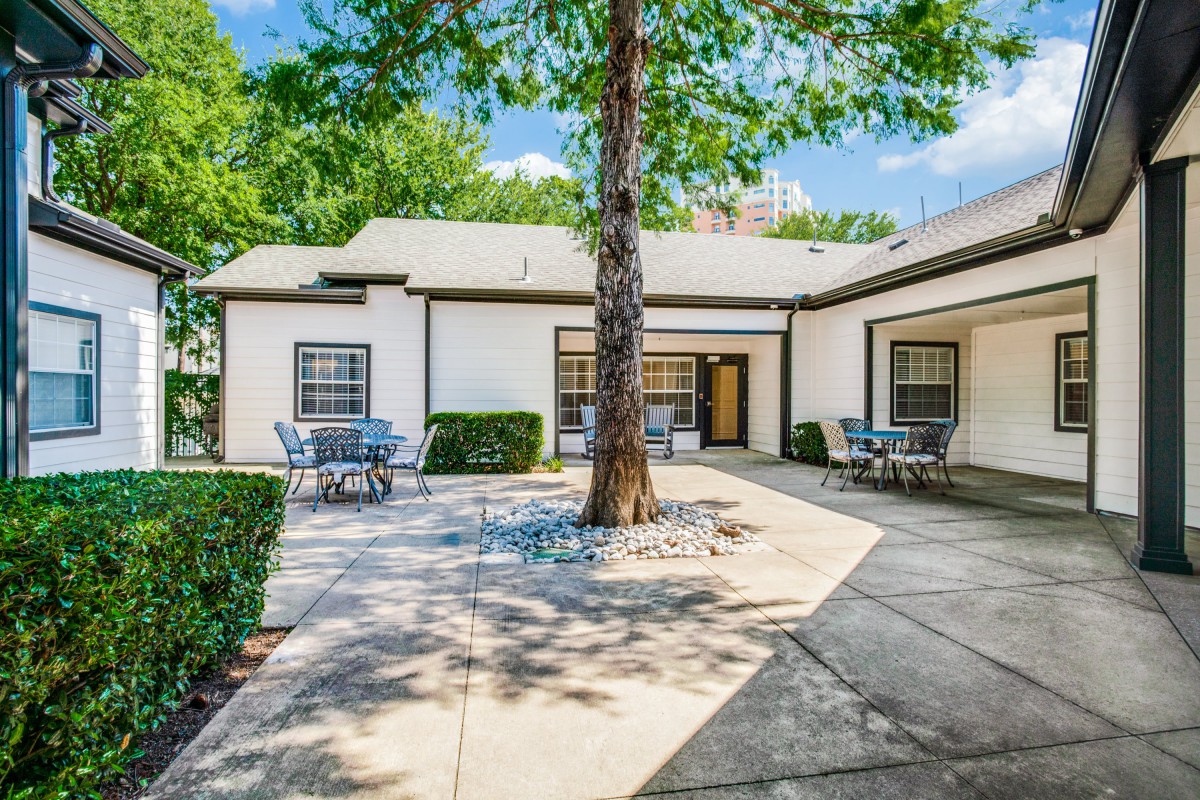 A tranquil courtyard featuring a large tree, surrounded by a building with tables and chairs under a clear sky.
