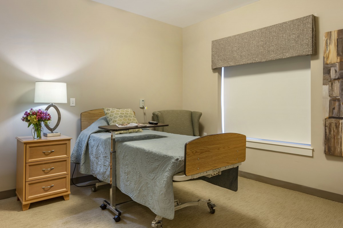 A clean and simple hospital room with a single bed, bedside table, and chair.
