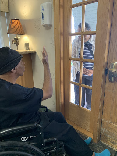 An elderly person in a wheelchair gestures towards a visitor through a glass door.