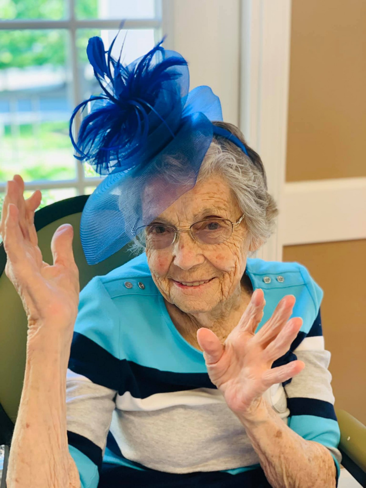 Elderly woman wearing a blue hat and glasses, smiling indoors.
