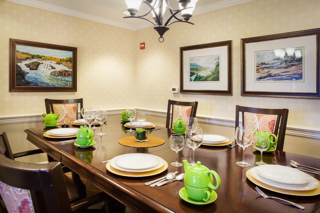 A neatly set dining table with green teapots and decorative plates in a warmly lit room featuring artwork on the walls.