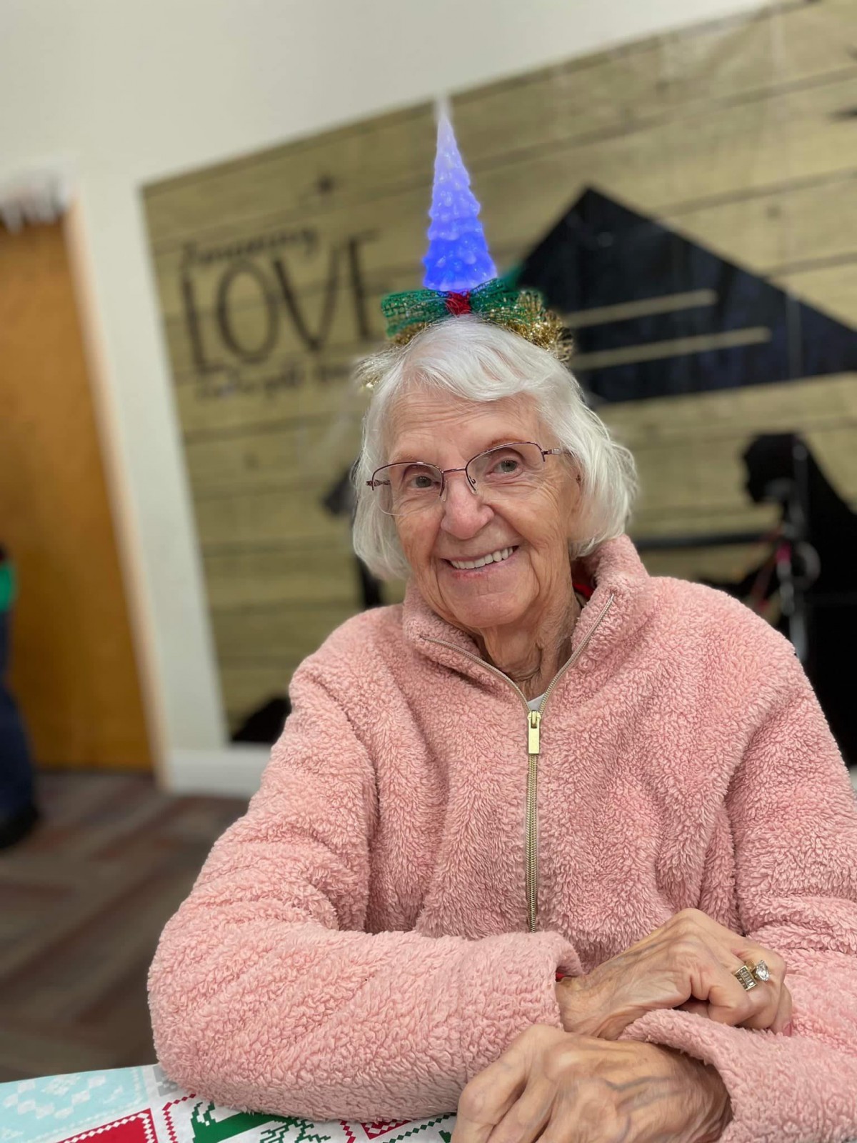 An elderly woman wearing a birthday hat and a pink sweater smiles warmly indoors.