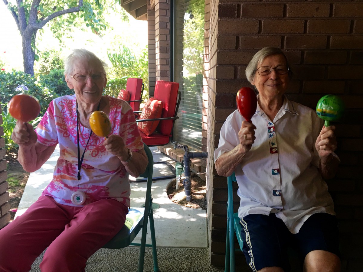 Two elderly women sitting together outdoors, smiling and holding maracas.