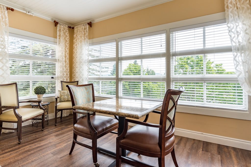 A well-lit dining area with a table, chairs, and large windows featuring blinds and curtains.