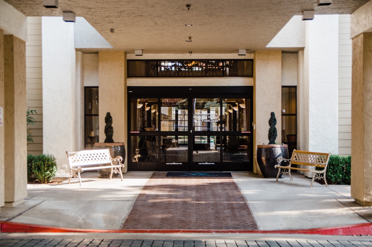 The entrance of a building with glass doors, flanked by benches and potted plants.