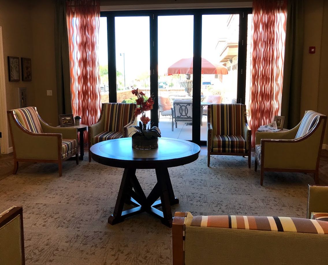 A cozy living room with a round table surrounded by chairs, featuring large windows and patterned curtains.