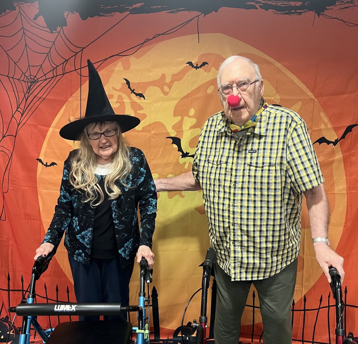 An older couple dressed up for Halloween, one as a witch and the other with a clown nose, posing in front of a festive backdrop.