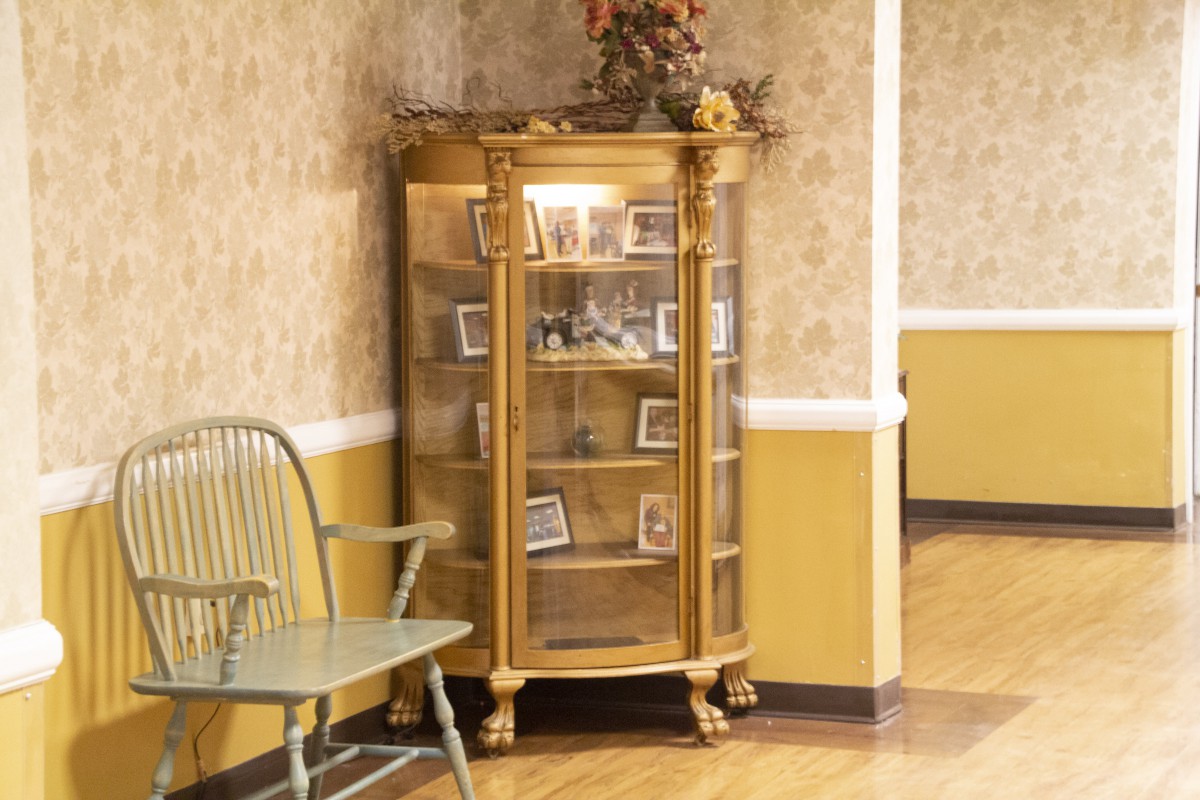 A vintage curio cabinet filled with photographs, placed against a wallpapered wall next to a chair.
