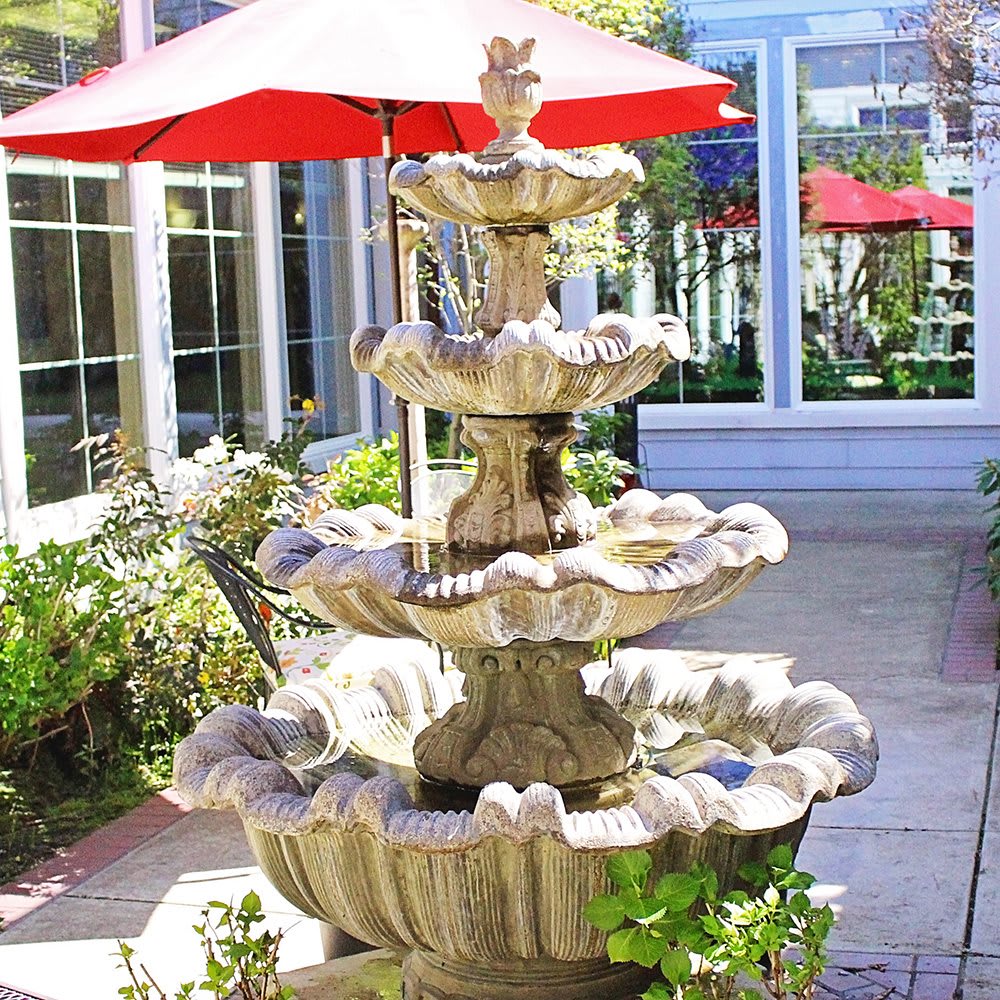 A decorative multi-tiered fountain in a sunlit courtyard with patio umbrellas.