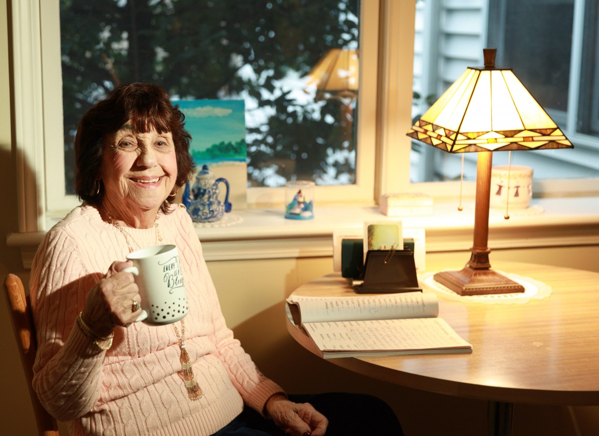 A woman sitting at a desk holding a coffee mug, surrounded by a cozy atmosphere with a lamp and books.