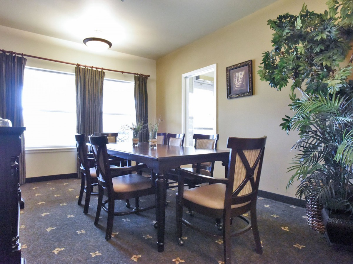 A well-lit dining room featuring a table with six chairs, large windows with curtains, and a patterned carpet.