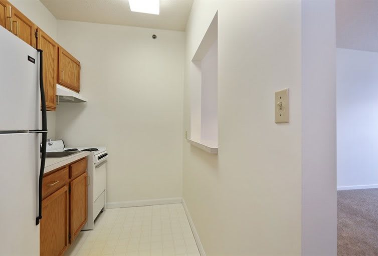 A compact apartment kitchen with white walls and wooden cabinets, featuring a stove, sink, and dishwasher.