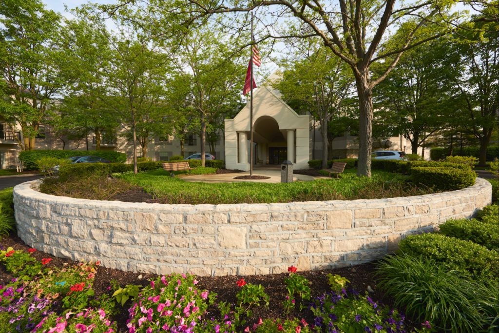 A landscaped park area with a stone wall, vibrant flowers, and trees, featuring a small building adorned with a flag.