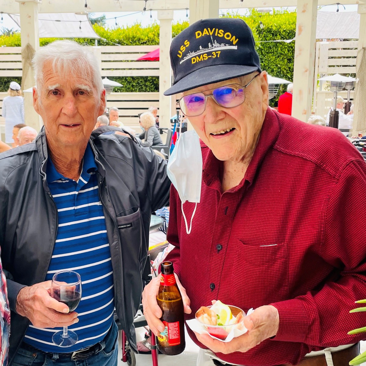 Two elderly men enjoying a social gathering outdoors, holding drinks and wearing casual attire.