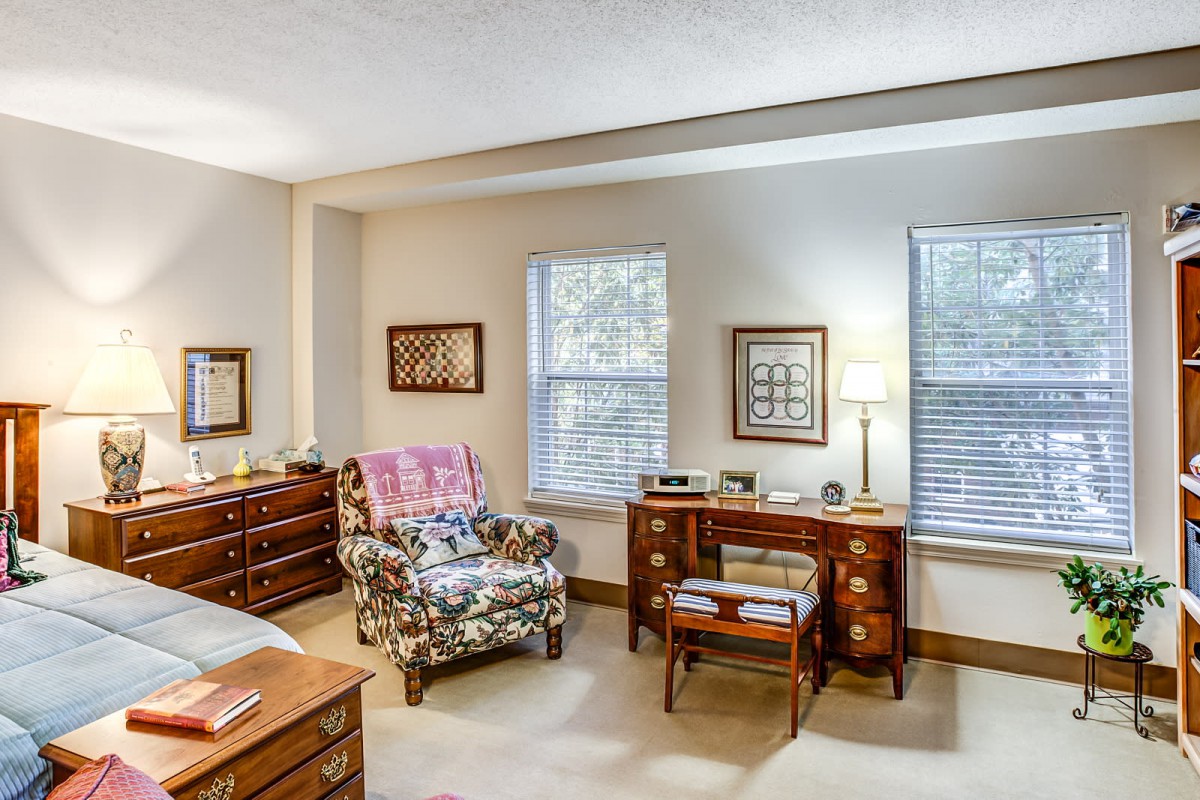 A cozy bedroom interior featuring a floral armchair, wooden desk, and two windows with shades.