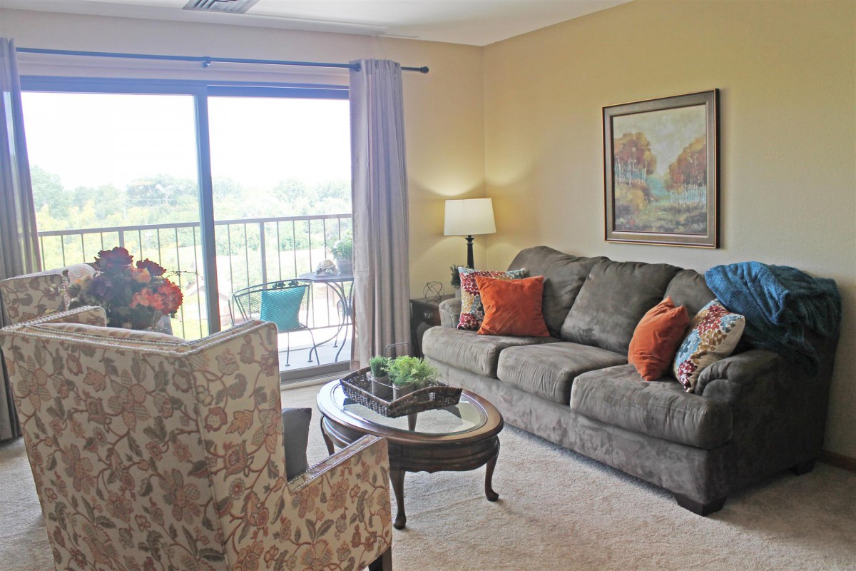A cozy living room with a grey sofa and patterned armchair, centered around a wooden coffee table. The space is enhanced by colorful cushions, a large painting on the wall, and natural light streaming through sliding glass doors leading to a balcony.
