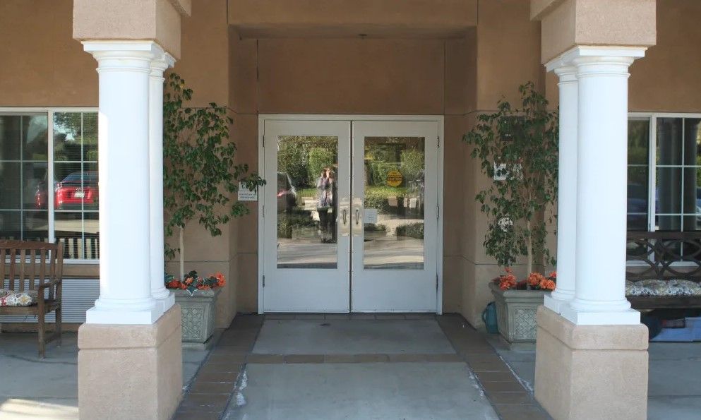 A building entrance with double glass doors, flanked by plants in pots, reflecting the outdoors.