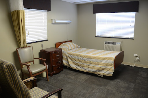 A simple room featuring a bed, chairs, and windows with blinds.