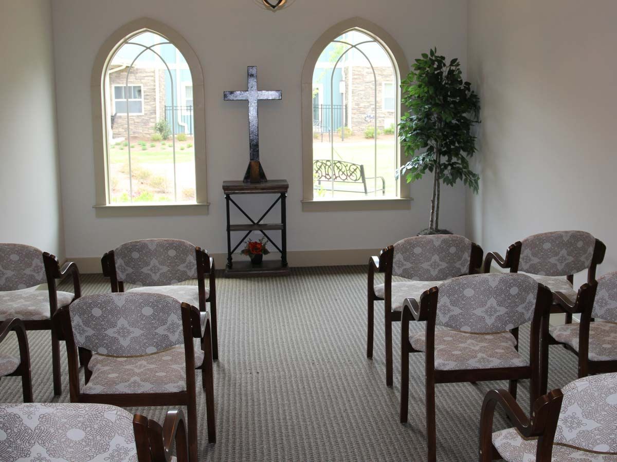A small chapel interior with rows of chairs, a cross at the front, and arched windows.