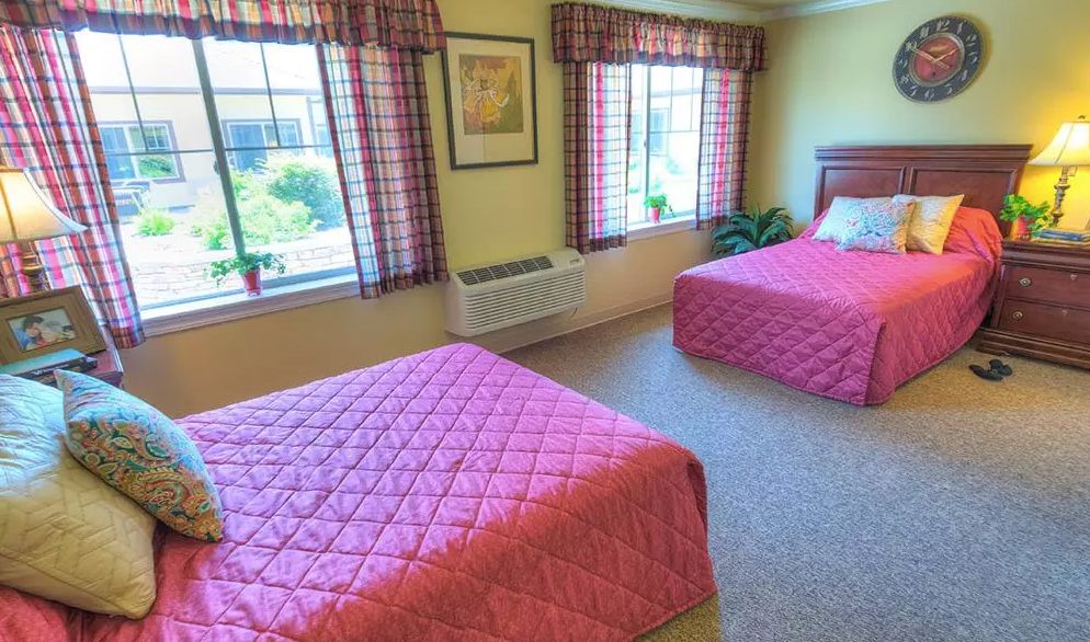 A cozy hotel room with twin beds and pink bedspreads, featuring large windows with checkered curtains.
