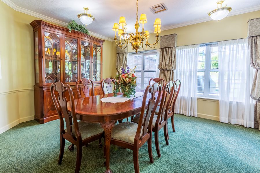 A traditional dining room with a large wooden table, elegant chandelier, and a carpeted floor.