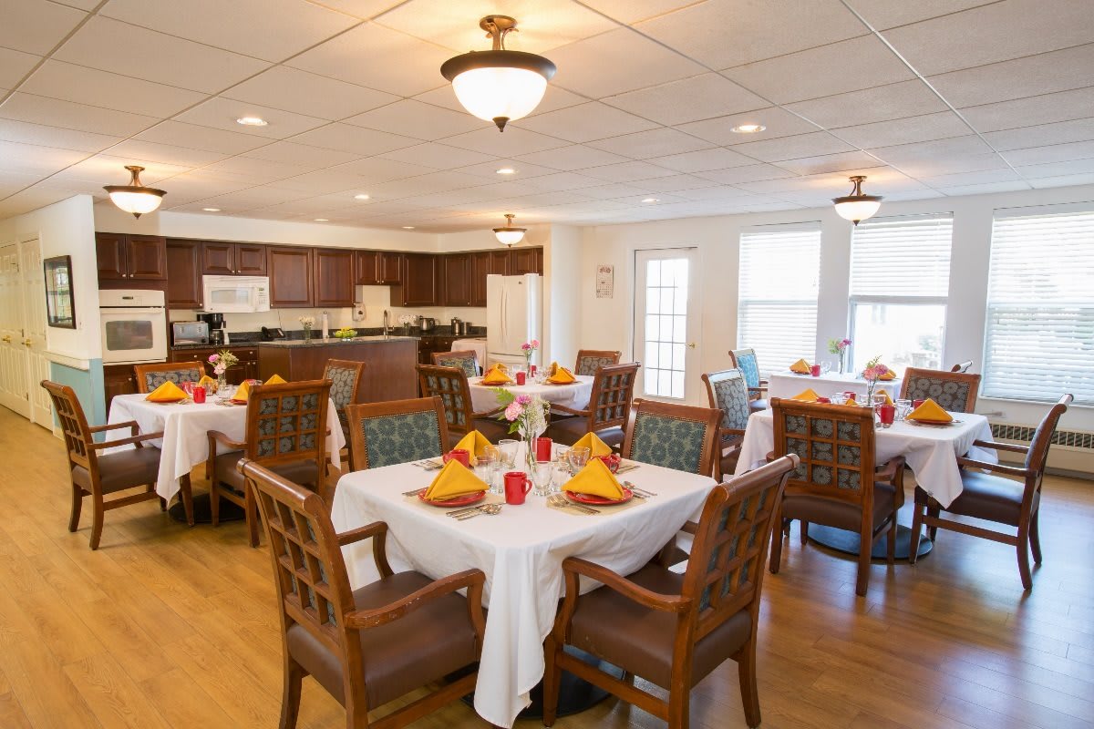 A well-lit dining room with several tables and chairs, featuring a kitchen area in the background.