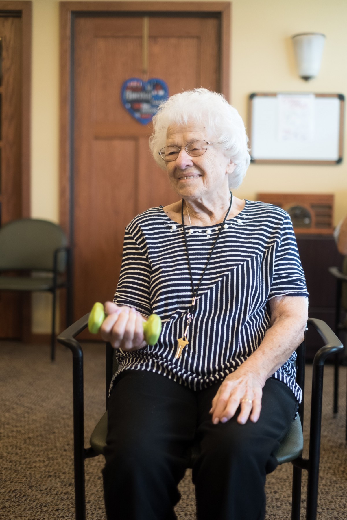 An elderly woman exercises with a dumbbell while seated indoors.