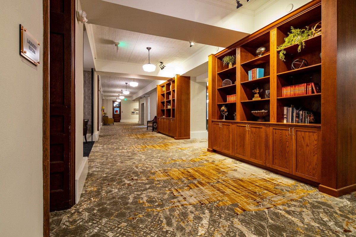 A well-lit hotel hallway with decorative carpet and wooden bookshelves.