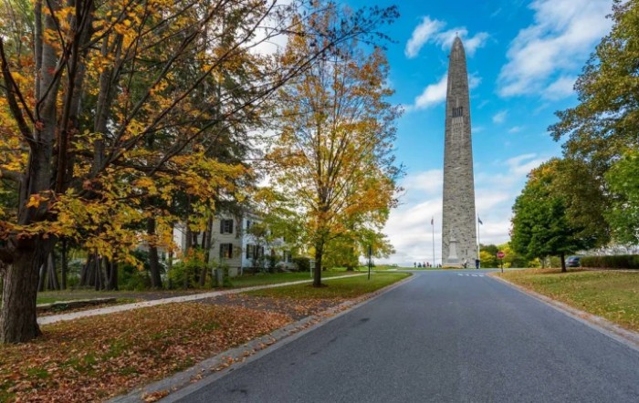 An obelisk towers over a tree-lined road with autumn foliage under a partly cloudy sky.