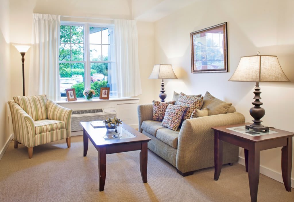 A cozy living room with a sofa, patterned armchair, and coffee table, featuring soft lighting and a large window with a scenic view.