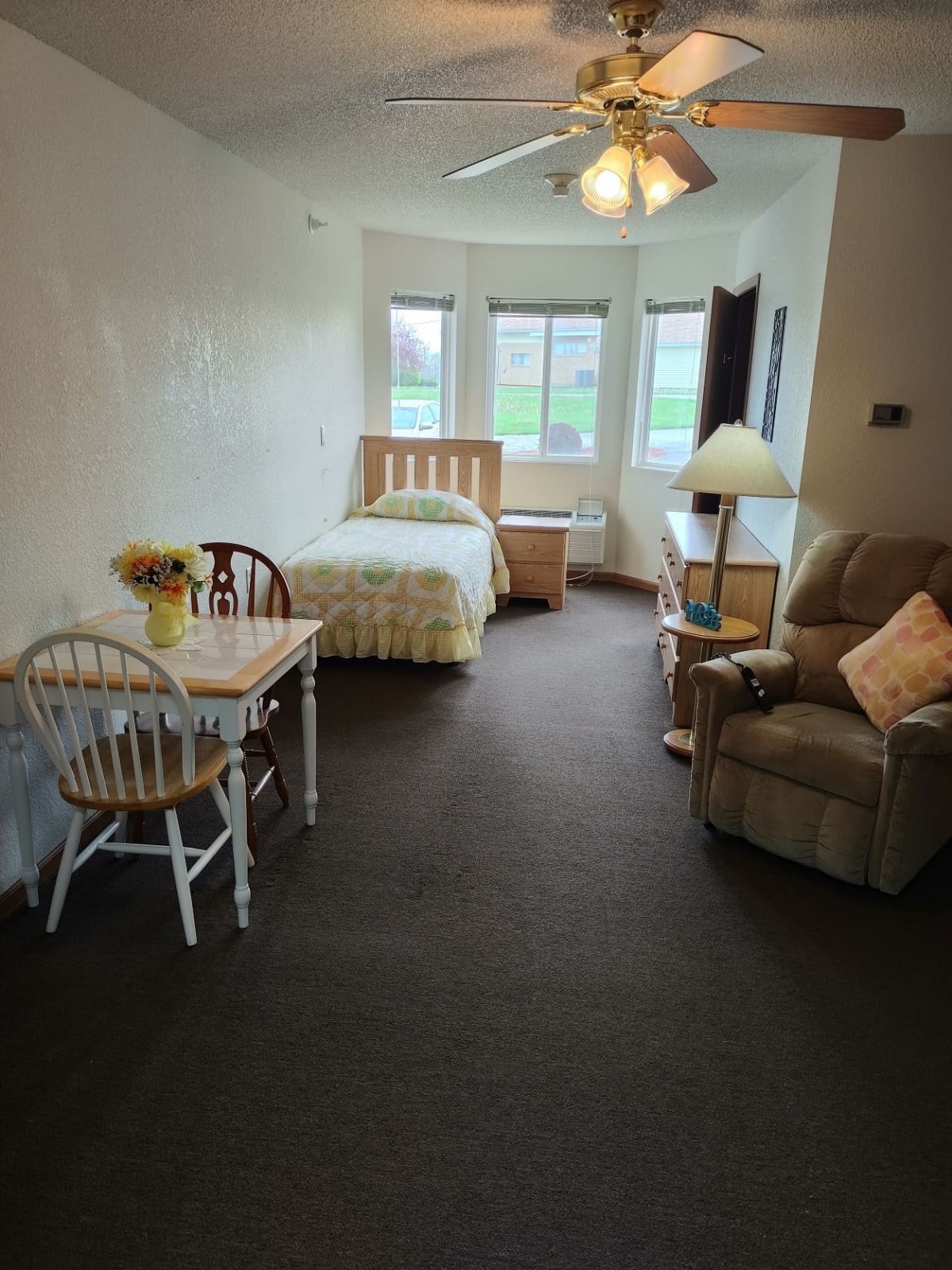 A cozy bedroom with a bed, table and chairs, reclining chair, lamp, and large windows.
