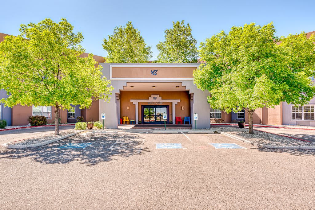 A spacious building entrance surrounded by lush trees and a parking lot under a clear blue sky.