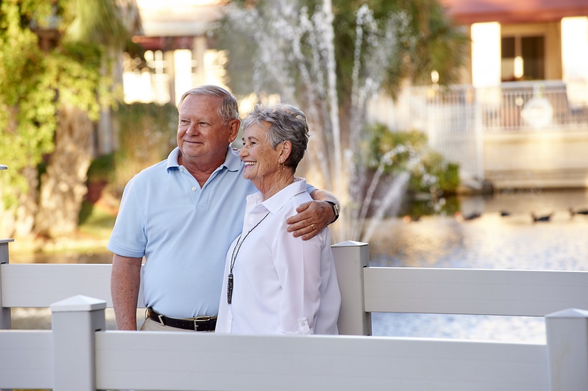 An elderly couple enjoys a sunny day by the fountain in a park.